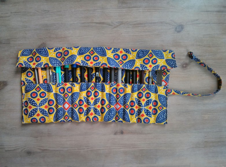 A yellow, red and blue repeat print pencil roll, fully unrolled and photographed from above on a wooden background. There are several pens, a metal rule and a mechanical pencil inside.